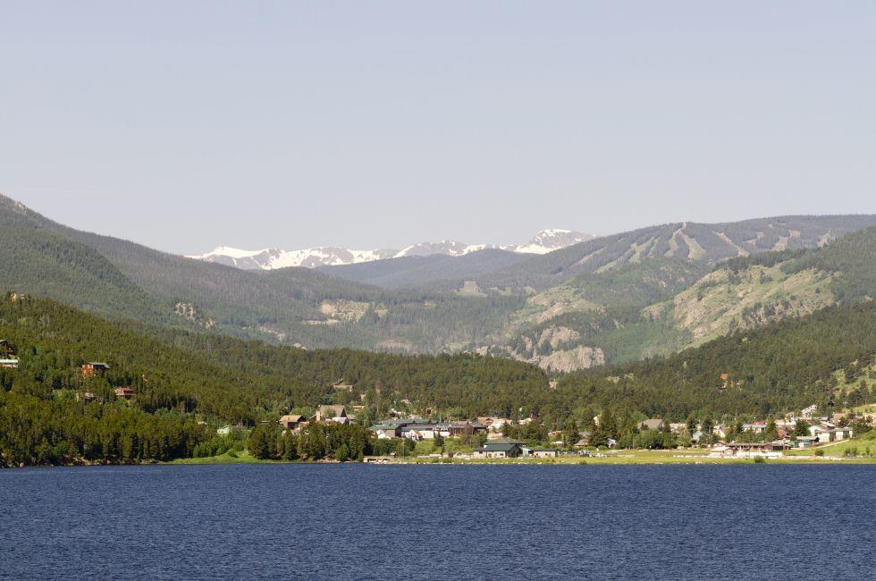 Waters of Barker Meadow Reservoir are foreground to town of Nederland in Colorado. Summer image shows unmelted snow in Indian Peaks Wilderness behind town. Evergreens cover slopes of hills. Horizontal image with copy space in pale blue sky above rural gate