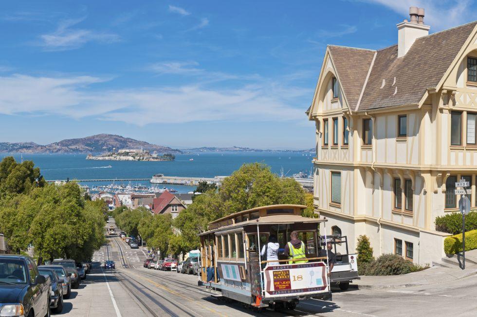 Tourists and locals riding the historic cable car trolley down a steep North Beach hill towards the blue ocean bay, harbor marina and Alcatraz beyond. Composite panoramic image created from four contemporaneous sequential photographs.