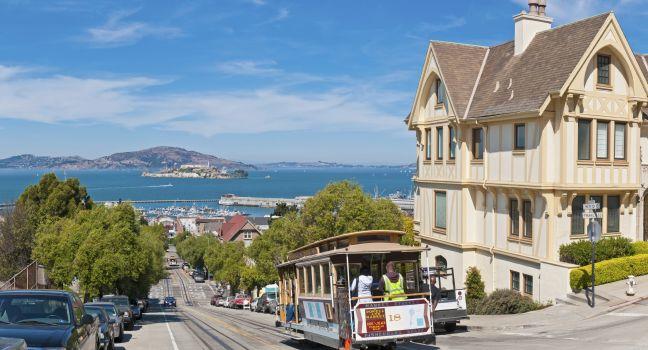 Tourists and locals riding the historic cable car trolley down a steep North Beach hill towards the blue ocean bay, harbor marina and Alcatraz beyond. Composite panoramic image created from four contemporaneous sequential photographs.