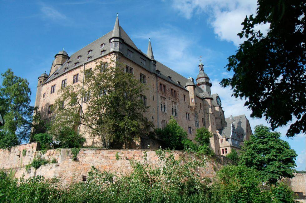 Marburg Castle in Germany on a clear day with clouds and a view of the towers, walls, clock and surrounding trees.
