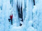 Athletic man in red coat with orange helmet and gold ice tools climbing a large blue wall of ice at the Ouray Ice Park